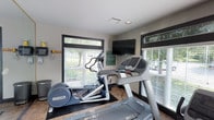 This single-floor fitness center is designed to provide a functional and comfortable environment for exercise. The space is outfitted with professional-grade Precor cardio equipment, including a treadmill and an elliptical trainer, both positioned to take advantage of the room's natural light. Large windows with horizontal blinds line the walls, offering views of the surrounding greenery outside and creating a bright, open atmosphere that makes workouts more enjoyable. A wall-mounted flat-screen television is prominently placed, allowing users to stay entertained during their sessions. The room features tile flooring with rubber exercise mats beneath the equipment for added safety and stability. Recessed ceiling lighting supplements the natural light, ensuring the space remains well-lit at all times. The neutral-toned walls and clean, uncluttered layout contribute to a focused workout environment. Informational signage is posted on the walls, providing guidelines for use of the facility. Overall, the fitness center is compact yet thoughtfully arranged, making efficient use of its footprint to deliver a practical and well-appointed amenity space.