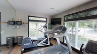 This single-floor fitness center is designed to provide a functional and comfortable environment for exercise. The space is outfitted with professional-grade Precor cardio equipment, including a treadmill and an elliptical trainer, both positioned to take advantage of the room's natural light. Large windows with horizontal blinds line the walls, offering views of the surrounding greenery outside and creating a bright, open atmosphere that makes workouts more enjoyable. A wall-mounted flat-screen television is prominently placed, allowing users to stay entertained during their sessions. The room features tile flooring with rubber exercise mats beneath the equipment for added safety and stability. Recessed ceiling lighting supplements the natural light, ensuring the space remains well-lit at all times. The neutral-toned walls and clean, uncluttered layout contribute to a focused workout environment. Informational signage is posted on the walls, providing guidelines for use of the facility. Overall, the fitness center is compact yet thoughtfully arranged, making efficient use of its footprint to deliver a practical and well-appointed amenity space.