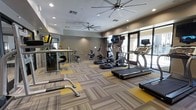 This single-floor fitness center is thoughtfully designed to provide a comprehensive exercise environment for users of all fitness levels. The space features a generous open layout with a patterned carpet tile floor accented by bold yellow geometric insets, adding a modern, energetic feel to the room. The ceiling is finished with recessed lighting and large industrial-style ceiling fans, ensuring the space remains well-lit and well-ventilated throughout workouts.

The cardio section is prominently featured and includes multiple treadmills, elliptical machines, and a stationary bike, several of which are equipped with personal viewing screens for an enhanced workout experience. On the strength and functional training side, the space offers a cable machine system, free weights, and medicine balls, providing a well-rounded selection of equipment.

Large glass doors and windows along one wall allow for ample natural light to flood the interior while offering views to an outdoor area, creating an open and inviting atmosphere. The neutral gray and taupe wall tones complement the modern equipment and flooring. Full-length mirrors are positioned strategically to maximize the sense of space and support proper form during exercise. Overall, this fitness center combines functionality with contemporary design to deliver a motivating and efficient workout environment.