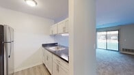 This single-story residence offers a practical and well-organized layout with clearly defined living spaces. The kitchen is a standout feature, designed in a galley style with white cabinetry fitted with brushed nickel hardware, dark countertops, and light wood-look vinyl flooring. Stainless steel appliances—including a refrigerator, electric range, and over-the-range microwave—provide a modern, cohesive look. A single-basin sink with under-cabinet lighting adds functionality to the workspace. The kitchen opens toward the main living area via a half-wall partition, promoting a connected feel between spaces.

The living area is generously proportioned, finished with neutral-toned carpet and a ceiling fan overhead. A large sliding glass door fitted with vertical blinds allows natural light to fill the room and provides outdoor access. Adjacent to the living space, a hallway connects to additional rooms, with multiple closet doors visible along the corridor.

The bedrooms are carpeted and finished in soft neutral tones, each receiving natural light through windows dressed with vertical blinds. One bedroom features a spacious walk-in closet with a distinctive accent-colored interior. The overall aesthetic throughout is clean, minimal, and move-in ready, with consistent neutral finishes across all spaces.