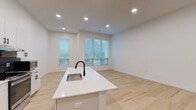 Located at 1101–1119 E 66th St in Kansas City, Missouri, this single-floor unit offers a clean, contemporary design centered around an open-concept living and kitchen area. Light hardwood flooring flows consistently throughout the space, contributing to a bright and cohesive aesthetic.

The kitchen is a standout feature, anchored by a large white quartz island with an integrated sink and matte black faucet. Crisp white cabinetry with black hardware extends to the ceiling, providing ample storage, while a bold dark tile backsplash adds visual contrast. Stainless steel appliances — including a French door refrigerator, gas range, and built-in microwave — complete the modern culinary setup.

The open living area benefits from multiple windows fitted with horizontal blinds, allowing natural light to filter in while offering views of surrounding greenery. Recessed lighting throughout ensures the space remains well-lit at all times.

A bathroom rounds out the unit, finished with the same light hardwood-style flooring and featuring an accessible design with stainless steel grab bars, a walk-in shower with tile surround and curtain, and clean white walls. The unit also includes two dark-finish interior doors, adding a modern touch to the overall design. The layout is efficient and well-planned, making strong use of the available footprint.
