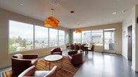 Located at 3261 SW Avalon Way in Seattle's West Seattle neighborhood, this rooftop lounge offers an elevated communal retreat with sweeping panoramic views of the Seattle skyline and surrounding neighborhood. The single-floor space is defined by floor-to-ceiling windows on multiple sides, flooding the interior with natural light while framing impressive cityscape vistas.

The lounge is thoughtfully divided into distinct functional zones. A casual seating area features rich brown leather barrel chairs arranged around a round coffee table, anchored by a vibrant multicolored striped area rug. Eye-catching sculptural orange pendant lights hang overhead, adding a bold, contemporary touch to the neutral palette of the space.

A dedicated dining area includes a live-edge wood table surrounded by leather chairs, positioned directly adjacent to large windows and a glass door that opens onto the outdoor rooftop terrace. The terrace, visible through the glazing, is equipped with outdoor lounge furniture, patio heaters, and a grill — ideal for gatherings and al fresco entertaining.

A built-in wall-mounted cabinet with decorative shelving provides functional storage and a design accent near the utility corridor. Two stainless steel elevators provide convenient access to the space. The overall ambiance is modern and welcoming, combining warm furnishings with clean architectural lines and outstanding views.