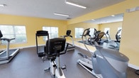 This fitness center offers a comprehensive workout environment designed to accommodate a range of exercise needs. The space is characterized by warm yellow walls, white trim, and durable rubber flooring, creating an energetic yet functional atmosphere. Fluorescent lighting runs along the ceiling, ensuring the room is consistently well-lit throughout.

The cardio section is well-stocked with professional-grade equipment, including True and Landice treadmills, an Octane elliptical trainer, and a True recumbent stationary bike. A large wall-mounted mirror spans one side of the room, visually expanding the space and allowing users to monitor their form during workouts.

For strength training, the facility includes a multi-station cable and weight machine, providing options for a full-body resistance workout. Small free weights are also available, adding versatility to training routines. A wall-mounted flat-screen television is positioned for visibility from multiple workout stations, offering entertainment during exercise sessions.

Multiple windows fitted with blinds allow natural light to filter in while maintaining privacy. Access to the room is provided through both a standard interior door and a glass-paned exterior door, contributing to an open and welcoming feel throughout the space.