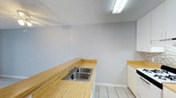 This single-floor space is designed with a clean, functional layout finished consistently in light-toned large-format tile flooring throughout. The kitchen serves as a practical hub, featuring warm wood-tone laminate countertops that extend generously along two walls, a stainless steel double-basin sink, and ample upper and lower white cabinetry. A gas range with an overhead exhaust fan sits against a neutral mosaic tile backsplash, adding a subtle textural detail. Overhead fluorescent lighting ensures the kitchen is well-illuminated.

The living area is open and simply appointed, painted in a soft gray tone with a ceiling fan overhead. A sliding glass door allows natural light to enter and provides access to an outdoor area. An adjacent room, likely a bedroom, features warm beige-toned walls, large windows fitted with vertical blinds for natural light control, and in-unit washer connections visible nearby.

The bathroom is straightforward and clean, equipped with a full-size tub and shower combination enclosed by a curved curtain rod, a toilet, and a pedestal-style sink, all finished in neutral tones. Throughout the space, white baseboards and neutral wall colors contribute to a cohesive, low-maintenance aesthetic.