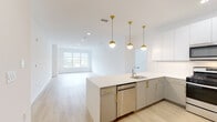 This single-floor unit showcases a clean, contemporary design defined by bright white walls, light hardwood flooring, and an abundance of natural light throughout. The open-concept kitchen and living area serves as the heart of the space, where a well-appointed kitchen commands attention with two-tone cabinetry — crisp white upper cabinets paired with warm gray lower cabinets — all accented by elegant brass hardware. A generous quartz countertop island with an undermount sink provides both workspace and a natural gathering point. Stainless steel appliances, including a gas range, microwave, and dishwasher, complete the functional kitchen. Pendant globe lights with gold fixtures hang above the island, adding a refined touch. The adjoining living area is spacious and airy, with large windows framing lush outdoor greenery and recessed lighting overhead. The bedrooms are comfortably scaled, each featuring light hardwood floors, neutral white walls, and windows that invite natural light. The bathrooms are thoughtfully designed, featuring wood-look tile flooring, a frameless glass-enclosed shower with gray tile surround, a white vanity with brass hardware, and a backlit LED mirror that adds a modern, spa-like quality to the space. The overall ambiance is fresh, minimal, and move-in ready.