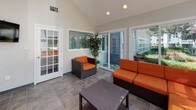 This inviting single-floor sunroom is defined by its standout architectural feature: a dramatic glass skylight ceiling that floods the interior with natural light throughout the day. The open, airy layout is grounded by durable slate-toned tile flooring, which extends seamlessly across the entire space and complements the clean, neutral wall palette.

The lounge area is furnished with a coordinated set of dark wicker furniture, including a generously sized sectional sofa and matching armchairs, all upholstered in vibrant orange cushions that add a warm, welcoming contrast to the cool-toned flooring and white walls. A low wicker coffee table anchors the seating arrangement, creating a comfortable and functional gathering area.

Vaulted ceilings rise toward the skylight, enhancing the sense of vertical space and openness. Multiple large sliding glass doors and windows line the walls, offering unobstructed views of the surrounding outdoor greenery and providing effortless indoor-outdoor connectivity to an adjacent patio area. A classic French door with glass pane detailing adds an elegant architectural accent.

A wall-mounted flat-screen television is positioned for comfortable viewing from the seating area, while recessed lighting supplements the abundant natural light. Lush potted plants are placed throughout, reinforcing the connection to the outdoors and adding a natural, refreshing ambiance to this versatile year-round retreat.