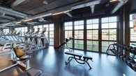 This expansive fitness center offers a comprehensive workout environment housed within a striking industrial-style space. The design features exposed concrete columns, high ceilings with visible ductwork and ventilation systems, and sleek linear lighting that illuminates the floor throughout. Large grid-pane windows line the perimeter, flooding the space with natural light and offering views of the surrounding outdoors.

The cardio zone is extensively equipped with a long row of treadmills, elliptical machines, rowing machines, and stationary bikes — including screen-equipped models for interactive training. Yellow accent markers on the light wood flooring define workout zones and add a modern visual contrast to the industrial aesthetic.

The strength training area features a full dumbbell rack with a wide range of weights, barbells, weight plates, and multiple cable and functional training machines, including Precor equipment. Adjustable benches and squat racks round out the free weights section. Wall-to-wall mirrors enhance the space and allow for form monitoring during lifts.

A dedicated open floor area with hardwood-style flooring accommodates flexibility and functional training, complete with stability balls and stretching accessories. The overall atmosphere is professional, modern, and well-organized, making this fitness center a thorough facility suited for a wide range of training needs.