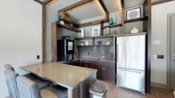 Located at 1414 Broadview Ave in Columbus, Ohio, this single-floor space offers a thoughtfully designed interior with a strong emphasis on modern aesthetics and functional design. The kitchen area serves as a standout feature, showcasing a sophisticated blend of dark wood cabinetry, a neutral-toned countertop island, and stainless steel appliances, including a full-sized refrigerator. A striking architectural detail draws the eye upward — an exposed wood beam framework with recessed lighting forms a distinctive ceiling accent that adds warmth and visual depth to the space. Open shelving integrated within the beam structure provides both decorative display space and practical storage, accented by curated décor pieces in muted and teal tones. A glass tile backsplash and undermount sink contribute to the clean, polished look of the kitchen workspace. A coffee station is conveniently positioned within the layout, enhancing the space's functionality. Light hardwood flooring runs throughout, complementing the overall warm yet contemporary palette. The single-floor layout promotes an open, accessible flow, making this space well-suited for both professional and social use.