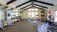 This fitness center is designed to offer a comprehensive workout environment within a single open-plan space. The room's most distinctive architectural feature is its vaulted ceiling, accented by bold dark wood exposed beams that run the length of the space, adding a striking visual contrast against the white ceiling. Recessed lighting and ceiling fans are integrated throughout, ensuring the room remains well-lit and comfortable during use.

Large windows, including a prominent multi-pane window along the front wall, allow generous natural light to fill the space, creating an open and inviting atmosphere. The warm neutral-toned walls complement the carpeted flooring, which spans the entire room.

The fitness center is divided into functional zones. The cardio area features multiple treadmills, elliptical trainers, and a rowing machine, each equipped with individual display screens. The strength training area includes a full dumbbell rack with a wide range of weights, a flat weight bench, and additional resistance equipment. Wall-mounted televisions are positioned throughout for entertainment during workouts. Full-length mirrors along one wall further enhance the sense of space while supporting proper form during exercise.