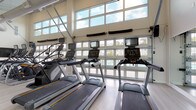 This single-story fitness center is designed with both functionality and openness in mind. The space is defined by soaring ceilings and an abundance of natural light, delivered through a striking combination of large transom windows positioned high along the walls and full-height garage-style glass panel doors that face an outdoor pool area. The light-toned hardwood-style flooring and crisp white walls contribute to a clean, modern aesthetic throughout.

The cardio zone is prominently featured along the windowed wall, housing a row of Matrix treadmills and elliptical cross-trainers, each equipped with individual touchscreen monitors. A ceiling fan overhead aids air circulation in this area. Adjacent to the cardio equipment, the strength and flexibility zone includes a rack of dumbbells, a resistance band station, a SmartMat core exercise board, and a weight bench, supported by wall-mounted mirrors and a flat-screen television.

A dedicated water station provides dual drinking fountains, ensuring convenience during workouts. Rolled towels are neatly arranged on a shelving unit, adding a thoughtful amenity. The overall ambiance is bright, spacious, and well-organized, creating an inviting environment suited for a comprehensive fitness routine.