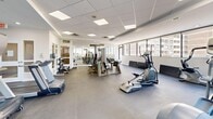 This fitness center offers a generously sized, single-floor exercise space designed to accommodate a comprehensive range of workout activities. The room features a drop ceiling with recessed panel lighting that evenly illuminates the entire area, complemented by abundant natural light streaming through large windows that line multiple walls. These windows frame expansive views of the surrounding urban skyline, creating an energizing atmosphere during workouts.

The flooring consists of durable large-format gray tiles, providing a clean, low-maintenance surface suited for high-traffic use. The space is divided functionally into distinct cardio and strength training zones. The cardio section includes multiple treadmills and elliptical trainers positioned along the windowed walls to maximize views and airflow. A stationary bike and stair climber round out the cardio offerings.

The strength training area houses a multi-station cable and weight machine, a dedicated free-weight rack stocked with a full range of dumbbells, and adjustable benches for varied exercises. A wall-mounted flat-screen television provides entertainment, and a sanitation station with wipes is conveniently positioned within the space. Accent walls in soft blue add a subtle visual contrast to the otherwise crisp white interior, contributing to the room's clean and motivating ambiance.