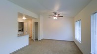 This single-story space features an open-concept living area finished with neutral wall-to-wall carpeting, creating a clean and versatile foundation suitable for a variety of furniture arrangements. A ceiling fan with an integrated light fixture is centrally mounted, providing both air circulation and ambient lighting to the main living space. Natural light enters through a window along the side wall, contributing to the bright and airy atmosphere of the room.

A partial wall with a raised breakfast bar counter separates the living area from the kitchen, maintaining visual connectivity between the two spaces. The kitchen area, visible from the living room, includes countertop space and cabinetry. Adjacent to the kitchen pass-through, a doorway leads further into the unit, while a dedicated closet with visible shelving provides convenient built-in storage. The smooth, light-colored walls and consistent neutral palette throughout lend the space a cohesive and understated character. Overall, the layout is practical and well-organized, offering functional living and storage areas within a compact, single-level footprint.