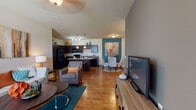 This well-appointed single-floor residence offers a thoughtfully designed open-concept layout that seamlessly connects the living, dining, and kitchen areas. Rich hardwood-style flooring flows throughout the main living spaces, complemented by vaulted ceilings that lend an airy, expansive feel. The living room is a welcoming focal point, featuring large windows that invite natural light and space for generous seating arrangements centered around a media console.

The kitchen is a standout feature, showcasing dark espresso cabinetry, granite-style countertops, black appliances, and a substantial island with bar seating—ideal for casual dining and entertaining. Pendant lighting above the island adds a refined touch. Adjacent to the kitchen, a dedicated dining area with a bold teal accent wall provides a vibrant setting for meals.

The primary bedroom offers a serene retreat with plush carpeting, large windows with ample natural light, and a neutral color palette. A second bedroom features a striking teal accent wall paired with rich, bold textiles, creating a cozy and colorful atmosphere. Both bathrooms are finished with hardwood-style flooring, dark vanity cabinetry, and shower-tub combinations. A dedicated in-unit laundry room with side-by-side washer and dryer adds everyday convenience to this well-rounded residence.