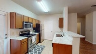 This single-floor residence offers a thoughtfully designed layout with a blend of practical features and comfortable living spaces. The kitchen is a functional centerpiece, outfitted with warm honey-toned cabinetry, light granite-style countertops, stainless steel appliances including a range with microwave above, and a refrigerator. Light tile flooring runs throughout the kitchen, which also benefits from a side door providing convenient exterior access. A peninsula with a sink adds workspace and visual separation between the kitchen and the adjoining dining area. The dining space is bright and welcoming, featuring large windows with blinds that allow ample natural light and a decorative chandelier that adds a touch of elegance overhead. The transition from tile to rich hardwood-style flooring marks the shift into the living and bedroom areas. A hallway connects the private rooms, each featuring ceiling fans, windows with blinds, and neutral wall tones. The bedrooms are carpeted for comfort and offer a quiet, restful atmosphere. The bathroom includes a generously sized soaking tub with tiled surround and built-in shelving niches, providing a relaxing retreat. Throughout, clean white trim and neutral paint tones create a cohesive and inviting ambiance.
