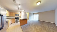 This single-floor residence offers a thoughtfully designed open-concept layout that seamlessly connects the kitchen and living areas, creating a bright and functional living environment. The kitchen is well-appointed with light maple cabinetry, dark countertops, and stainless steel appliances including a gas range and built-in microwave. A central kitchen island provides additional prep space and serves as a natural dividing point between the kitchen and the rest of the living area. Track lighting overhead ensures the kitchen is well-illuminated throughout the day. The transition from the kitchen's hardwood-style flooring to the plush carpeting of the living room marks the boundary of the open living space, which is generously sized and filled with a sense of openness. A large sliding glass door fitted with vertical blinds allows natural light to pour in and offers convenient access to the outdoors. Neutral wall tones and standard ceiling heights contribute to a clean, welcoming ambiance throughout. A private entry door is also visible, adding to the practical layout of the home. Overall, this space balances comfort and functionality in a cohesive, move-in ready design.