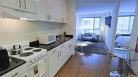 This single-floor unit features an open-concept layout that seamlessly connects a functional kitchen to a well-appointed living area. The kitchen is outfitted with crisp white cabinetry, dark stone countertops, and white appliances including an electric range and microwave, providing a clean, contemporary aesthetic. A built-in sink with a black surface adds a sleek contrast against the white lower and upper cabinets.

The living area extends naturally from the kitchen, anchored by warm hardwood-style flooring that runs throughout the space. A large floor-to-ceiling window at the far end floods the room with natural light, creating an airy, open ambiance. The living space comfortably accommodates a sofa set arranged around a coffee table, complemented by a textured area rug that adds depth and warmth. A wall-mounted flat-screen television and slim floor lamps contribute to the modern, minimalist style. The overall design balances practicality with contemporary style, making efficient use of the available space while maintaining a cohesive and inviting atmosphere throughout.