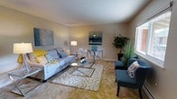 This single-floor residence offers a warm and inviting atmosphere, highlighted by a well-appointed living area that serves as the heart of the home. The living room features plush wall-to-wall carpeting in a soft golden-beige tone, creating a cozy foundation underfoot. Neutral-painted walls provide a versatile backdrop, while a textured popcorn ceiling adds classic character to the space.

The room is generously proportioned, offering ample space for comfortable seating arrangements. A large, light-toned sofa anchors the space, accompanied by stylish accent chairs upholstered in a complementary slate blue, giving the room a balanced, curated feel. A geometric chrome-framed coffee table rests atop a patterned area rug, adding a contemporary touch to the overall design.

Natural light filters in through a window fitted with a Roman shade, brightening the interior alongside warm ambient lighting from a floor lamp. Decorative wall art in cool oceanic blues adds visual interest and ties together the home's cohesive color palette of blues, grays, and warm neutrals. A console table with matching decorative accents and lush indoor greenery further enhance the space, creating an atmosphere that feels both polished and livable.