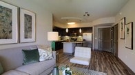 This single-floor residence showcases a thoughtfully designed open-concept layout that seamlessly connects the living, dining, and kitchen areas into one cohesive space. The living area provides a comfortable setting with neutral-toned walls and warm wood-style flooring that extends throughout the unit, creating a consistent and inviting atmosphere.

The kitchen is a standout feature, outfitted with dark espresso cabinetry that reaches toward the ceiling, offering generous storage. A mosaic tile backsplash in muted greens and grays adds visual texture behind the range and countertops. Stainless steel appliances, including a refrigerator, microwave, and dishwasher, provide a polished, modern aesthetic. A curved white breakfast bar island serves as both a functional food preparation surface and a casual dining space, accompanied by contemporary bar seating.

Pendant lighting in elongated cylindrical forms hangs above the island, complementing the overall flush ceiling fixtures to provide layered illumination throughout the space. The open sightlines between the kitchen and living area enhance the sense of spaciousness, making the layout feel airy and well-connected. The combination of dark cabinetry, light countertops, and wood flooring gives the space a balanced, contemporary character.