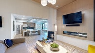 This thoughtfully designed single-floor communal space blends contemporary aesthetics with functional versatility. The living area commands attention with its soaring ceilings, warm wood-paneled accents, and a dramatic light wood ceiling feature adorned with a cluster of globe pendant lights. A sleek wall-mounted fireplace is framed by a natural wood surround, complemented by a large flat-screen television above. Seating options are varied and intentional — a plush gray sofa, vibrant yellow accent chairs, a navy leather chair, and a built-in banquette with small round orange-legged tables create distinct zones for relaxation and socializing. A deep green feature wall fitted with circular illuminated sconces adds a bold, artistic focal point. Light hardwood flooring runs throughout, anchored by a multicolored geometric area rug in the lounge zone. A dedicated game area houses a full-length shuffleboard table. The kitchen area transitions seamlessly from the lounge, featuring a large white island with an integrated wood bar, a herringbone wood backsplash, bold navy tile accents, stainless steel appliances including a full-sized refrigerator, and exposed industrial ductwork overhead that adds an urban, loft-inspired character. The entire space is bright, airy, and designed for community engagement.