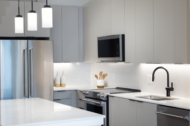 Cocina Signature Collection con electrodomésticos de acero inoxidable, gabinetes de color gris claro, encimeras y protector contra salpicaduras de cuarzo marmolado, iluminación debajo de los gabinetes e iluminación colgante. - Avalon at Becker Farm