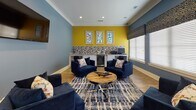 This thoughtfully designed single-floor communal space offers a welcoming and sophisticated atmosphere through a cohesive blend of bold color and refined furnishings. The interior is defined by rich blue walls that carry consistently throughout, complemented by warm hardwood flooring that unifies the various areas seamlessly.

The main lounge area is generously sized and centered around plush navy blue seating arrangements accented with floral throw pillows, grouped around a distinctive round wooden coffee table atop a patterned area rug. A wall-mounted flat-screen television serves as a focal point on one end, while large windows fitted with white horizontal blinds and decorative fabric valances allow natural light to filter in softly.

Elegant white French doors with glass pane inserts connect the lounge to an adjoining gathering room, which features soaring ceilings with exposed white beams and a statement chandelier. This secondary space is furnished with vibrant mustard yellow accent chairs, a neutral sofa, and curated artwork, creating an inviting conversational setting.

A beverage bar area anchors one wall, highlighted by a bold yellow accent wall, mosaic tile backsplash, under-counter refrigeration, and framed geometric artwork above. Recessed lighting throughout ensures consistent, ambient illumination. Decorative shelving and layered accessories complete the polished, community-oriented aesthetic of this well-appointed space.