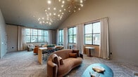 Located at 6615 Cerris Dr in Charlotte, NC, this thoughtfully designed community lounge space offers a sophisticated and welcoming atmosphere across a single floor. The layout includes a spacious living room and a connecting hallway, together forming a well-appointed gathering area.

The living room is the centerpiece of the space, anchored by dramatic vaulted ceilings adorned with a striking starburst chandelier featuring multiple brass-tipped arms — an immediately eye-catching focal point. Large black-framed windows dressed with floor-to-ceiling linen curtains flood the interior with natural light and offer views of the surrounding neighborhood.

The room is furnished with a mix of curved sofas in warm cognac tones, paired with soft grey accent chairs and a teal oval coffee table, creating an inviting and eclectic aesthetic. Patterned carpeting with an organic motif adds texture and warmth underfoot. A dedicated dining and collaboration area features booth-style seating tucked beneath elegant arched alcoves, each illuminated by brass pendant lights — ideal for small gatherings or focused work.

A sculptural wooden room divider with oval cutouts elegantly separates zones within the space, while wall-mounted mirrors amplify depth and light. The hallway connects seamlessly to the main area, reinforcing the space's open, cohesive flow. Overall, the design balances modern sophistication with approachable comfort.