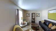 This single-floor residence offers a comfortable and thoughtfully designed living space that blends functionality with style. The living room serves as the heart of the home, featuring neutral warm-toned walls that create a welcoming ambiance. Natural light filters in through a large window dressed with patterned curtains, brightening the room and highlighting the carefully curated furnishings. The space is anchored by a plush sofa and coordinating armchairs, complemented by a round wooden coffee table that adds a natural, organic element to the layout. A patterned area rug defines the seating area and adds visual interest underfoot. A dark wood open shelving unit displays decorative accents, while a pair of framed artwork pieces on the wall contribute a touch of color and personality to the neutral palette. Accent pillows in warm gold and muted tones tie the color scheme together cohesively. The vaulted ceiling adds a sense of openness and volume to the main living area, enhancing the overall sense of space. The layout flows naturally, making the home feel both practical and inviting — ideal for everyday living and relaxed entertaining.