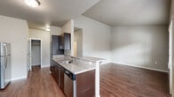This single-floor, 4-bedroom, 1-bathroom unit offers a practical and modern layout designed to accommodate multiple occupants comfortably. The open-concept kitchen and living area serves as the heart of the unit, featuring rich dark cabinetry, granite-style countertops, a peninsula with a built-in sink, and a full suite of stainless steel appliances including an electric range, microwave, refrigerator, and dishwasher. Hardwood-style flooring flows throughout the main living areas, complementing the neutral gray walls and clean-lined aesthetic. The living area receives natural light through a large window and provides access to an outdoor balcony via a glass-paneled door. The four bedrooms are carpeted for comfort, with each room offering neutral tones and natural light from individual windows. One bedroom notably includes access to an in-unit laundry area with a washer and dryer. The shared full bathroom is finished with tile flooring, a granite-topped vanity with a dark cabinet base, a toilet, and a walk-in shower. A hallway connects the bedrooms and bathroom, providing a logical and efficient flow throughout the unit. The overall design emphasizes functionality with consistent, contemporary finishes.