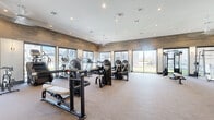 This well-appointed fitness center offers a generously sized, single-floor layout designed to accommodate a comprehensive range of workout activities. The main exercise area is outfitted with an extensive selection of equipment, including selectorized strength machines, cable systems, free weights, dumbbells, medicine balls, and multiple cardio machines such as treadmills and ellipticals. The open floor plan allows for ample circulation between stations, creating a comfortable and functional training environment.

The interior design balances modern aesthetics with a warm atmosphere. Wood-plank accent walls extend along the upper perimeter, complementing the neutral-toned carpet flooring throughout. Recessed lighting in the white ceiling provides consistent, even illumination across the entire space. Large sliding glass doors and expansive windows line multiple walls, flooding the fitness area with natural light and offering unobstructed views of the surrounding outdoor grounds.

A dedicated free weights and functional training zone features floor-to-ceiling mirrors, maximizing the sense of space. A separate entry corridor with glass-partition walls, tile flooring, and contemporary artwork provides a polished transition into the facility. Direct exterior access through glass doors further enhances the open, connected feel of this well-equipped fitness space.