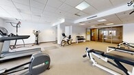 Located at 7301 Rue Beaubien E in Anjou, Québec, this single-floor amenity space provides residents with a functional and practical set of shared facilities. The layout encompasses a lobby/hallway area, a dedicated gym, and additional support rooms.

The entry lobby sets a welcoming tone with tile flooring in a herringbone pattern, recessed lighting within a drop-panel ceiling, and warm neutral wall tones. A black leather loveseat and a wooden coffee table offer a comfortable waiting area, while a built-in wall of individual mailboxes serves as a practical mail collection point. An elevator and clearly marked exits ensure easy access throughout the building.

The fitness room is generously proportioned and well-lit by both recessed and fluorescent panel lighting. Carpeted flooring spans the workout area, which is outfitted with a broad selection of equipment including a treadmill, elliptical trainer, stationary bikes, a multi-station cable machine, weight benches, free weights, and a Precor rowing machine. Windows along one wall introduce natural light, enhancing the open, airy atmosphere of the space. The clean white walls and uncluttered layout make for a focused and functional workout environment suitable for a variety of fitness routines.