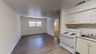Located at 2301 Old Bainbridge Rd in Tallahassee, Florida, this single-floor unit offers a straightforward, functional layout designed for comfortable everyday living. The main living and kitchen area is an open, light-filled space featuring durable wood-style laminate flooring throughout and a textured ceiling. Windows along the main wall allow natural light to brighten the room, creating an airy atmosphere. The kitchen is positioned adjacent to the living area and is equipped with white cabinetry, an electric range, and an overhead ventilation hood, making efficient use of the available space. A doorway from the living area leads into the bedroom, which features plush wall-to-wall carpeting and a ceiling fan with integrated lighting, providing both comfort and airflow. A natural light source from a window looks out toward greenery. The bedroom includes access to a generously sized walk-in closet outfitted with dual shelving rods on both sides, offering ample storage capacity. The bathroom presents a clean, functional design with wood-style flooring, a full soaking tub and shower combination accented by warm golden mosaic tile on the surround, and standard white fixtures throughout. Clean white walls are consistent across the entire unit, lending a bright, neutral backdrop throughout this practical Tallahassee residence.