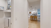 This single-floor residence is designed with a clean, contemporary aesthetic highlighted by light wood-style flooring, white walls, and recessed lighting that runs consistently throughout the space. The open-concept kitchen serves as a central feature, outfitted with white shaker-style cabinetry, granite countertops, a white subway tile backsplash, and a full suite of stainless steel appliances including a French door refrigerator, range, microwave, and dishwasher. A central island with seating provides additional workspace and casual dining. Pendant lights hang above the island, adding a polished touch. The kitchen opens directly into the living area, which benefits from large windows fitted with blinds and sheer curtains, allowing ample natural light. A ceiling fan adds comfort to the living space, which also includes access to an exterior door. The bedrooms maintain the same light, airy palette, each featuring wood-style flooring and generously sized windows. The bathroom echoes the kitchen's finishes with granite countertops and white cabinetry. A dedicated laundry area with a full-size washer and dryer adds convenience to the unit's practical layout.