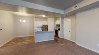 This single-floor residence offers a thoughtfully designed open-concept layout that seamlessly connects the kitchen and living areas. The kitchen is well-appointed with white shaker-style cabinetry, granite countertops, and a full suite of stainless steel appliances, including a refrigerator, electric range, and built-in microwave. A peninsula with an integrated sink and breakfast bar serves as both a functional workspace and a natural divider between the kitchen and the adjacent living space. A blue subway tile backsplash adds a subtle decorative accent to the kitchen. The living area features plush wall-to-wall carpeting, while the kitchen and entryway are finished with hardwood-style flooring, creating a clear and practical distinction between zones. Neutral gray walls and white trim throughout give the space a clean, contemporary feel. An elegant pendant chandelier anchors the dining area, complementing the recessed lighting in the kitchen. A solid white entry door provides a welcoming focal point near the transition to the main living space. Overall, the residence presents a modern, move-in-ready environment with a cohesive and functional design.