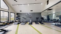 The fitness center at Prospect Pointe Apartments, located at 330 S New Prospect Rd in Jackson, NJ, is a thoughtfully designed exercise space that combines functionality with a modern aesthetic. The room is defined by its striking vaulted ceilings, which soar upward and are complemented by dramatic floor-to-ceiling windows along one wall. These large, black-framed windows flood the space with natural light and offer views of the outdoor pool area, creating an open and energizing atmosphere. Track lighting and ceiling fans are mounted overhead, ensuring both comfort and visibility throughout the room. The light-toned luxury vinyl flooring features distinctive green accent stripes that add a dynamic visual element to the space. A full wall of mirrors on one side visually expands the room and assists users during strength training. The fitness center is comprehensively equipped with a wide array of machines and free weights, including treadmills, ellipticals, stationary spin bikes with digital displays, a rowing machine, cable and weight machines, a full dumbbell rack, kettlebells, medicine balls, and adjustable benches. A textured accent wall displaying the Prospect Pointe branding adds a polished, branded finish to this well-appointed community amenity.