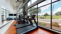 This fitness center is designed with both functionality and aesthetics in mind, offering a well-equipped workout environment within a contemporary architectural setting. The space is defined by soaring ceilings and expansive floor-to-ceiling windows framed in sleek black metal, which span the length of one wall and flood the interior with natural light while providing open views of surrounding greenery and outdoor scenery.

The vibrant orange rubber flooring anchors the space visually and provides a durable, high-performance surface suited to intensive exercise. Along the windowed wall, a row of Star Trac treadmills and elliptical machines are positioned to take full advantage of the outdoor views, creating an energizing cardio zone. Complementing these are air bikes and additional cardio equipment for varied training options.

The strength training area features a multi-function cable and power rack system, along with a well-organized dumbbell rack offering a broad range of weights. Wall-mounted flat-screen televisions are positioned throughout for entertainment and instructional use. Exposed ductwork running along the upper walls adds an industrial character to the otherwise clean, modern interior. A glass-paneled entrance leads into the space, contributing to its open and accessible feel.