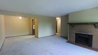 This single-story space centers around a generously sized living area finished with wall-to-wall carpet in a neutral tone, providing a clean and functional foundation. The room is defined by a classic brick fireplace positioned along one wall, complete with a simple wood mantle, serving as a natural focal point for the space. The walls are painted in a muted, earthy tone that complements the understated aesthetic throughout.

The open layout allows for clear sightlines to adjoining areas. A partially visible room to the left appears to be an additional living or utility space, accessible through a dark-trimmed doorway. A hallway visible toward the back of the space leads further into the home, suggesting additional rooms beyond. Overhead lighting provides ambient illumination throughout.

The absence of ornate detailing gives the space a straightforward, practical character. The combination of carpeted flooring, neutral walls, and brick fireplace creates a livable, cohesive environment. The single-floor layout offers convenient accessibility and ease of navigation throughout the home.