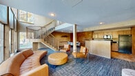 Located at Echo Park Circle in Burnsville, Minnesota, this single-floor community clubhouse offers a well-appointed gathering space designed for both relaxation and activity. The living room is a standout feature, characterized by soaring two-story windows that flood the area with natural light and provide views of the outdoor pool. Warm, richly textured faux-finish walls in amber and bronze tones lend an upscale ambiance throughout. The seating area is anchored by a curved tan leather sofa, a tufted ottoman, and modern accent chairs, all set atop a patterned blue carpet. A graceful curved staircase with metal railings adds architectural elegance to the space. The kitchen and dining area are outfitted with light maple cabinetry, dark granite countertops, a full suite of appliances, and a glass-top dining table with seating for six — ideal for community gatherings or events. The exercise room is well-equipped with cardio machines, a cable weight system, free weights, and a weight bench. Large French doors in the fitness area open to views of the pool, while full-length mirrors and a combination of rubber and tile flooring complete the practical, purpose-built layout.