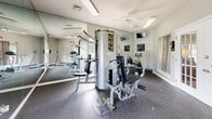 Located at 100 Willow Creek Dr in Mount Juliet, Tennessee, this fitness center offers a well-equipped exercise space designed to support a variety of workout routines. The room features durable speckled rubber flooring that provides both safety and comfort during exercise, while large windows along the walls invite ample natural light and offer views of the surrounding outdoor greenery. Full-length mirrors line multiple walls, enhancing the sense of space and allowing users to monitor their form during workouts. A ceiling fan keeps air circulating throughout the room, contributing to a comfortable environment. The equipment selection is comprehensive for the room's footprint, including a treadmill, recumbent bike, multi-station cable strength machine, and a weight bench, catering to both cardiovascular and resistance training needs. A wall-mounted television provides entertainment during workouts. The neutral gray walls and white trim give the space a clean, modern aesthetic. French-style doors connect the fitness center to an adjacent clubroom, and keypad-controlled access ensures secure entry. Posted fitness center rules and instructional signage round out the functional, community-oriented design of this exercise facility.