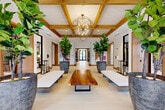 Located at 776 Riverside Dr in Coral Springs, Florida, this thoughtfully designed lobby and hallway space offers a refined and welcoming first impression. The single-floor layout comprises two hallways that together create a seamless and flowing passage through the space.

The main lobby commands attention with its exposed wood beam coffered ceiling, which adds warmth and architectural depth. A dramatic spherical brass chandelier serves as the centerpiece, casting a warm glow over the room. The star feature is a stunning live-edge wood slab table positioned along the central axis, flanked by sleek white upholstered benches that invite guests to pause and linger.

Oversized fiddle-leaf fig trees in large textured stone planters bring vibrant natural life to the space, reinforcing a resort-like ambiance. Vivid, colorful artwork displayed along the walls adds a curated, gallery-quality aesthetic. Light-toned tile flooring runs throughout, reflecting natural light and enhancing the airy feel.

The secondary hallway transitions into a more intimate passage clad in rich warm wood paneling, with black-framed glass doors and a coordinating chandelier. Floor-to-ceiling glass entries at both ends of the space frame outdoor views, flooding the interiors with abundant natural light and connecting the space visually to the lush Florida surroundings.