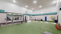 This single-story sports training facility is purpose-built for athletic performance and functional fitness. The main training area is dominated by a wide expanse of artificial turf, providing an ideal surface for agility drills, sled pulls, battle rope exercises, and speed training. The high ceilings amplify the sense of openness and accommodate a full range of dynamic movements without restriction. Recessed lighting throughout ensures bright, even illumination across the entire floor.

A row of large mirrors lines one wall, flanked by a dedicated strength zone featuring a cable machine, power racks, and a weight bench with barbells. An adjacent area with hardwood-style flooring provides space for machine-based strength equipment and rubber mat stations. Dumbbell and kettlebell racks are neatly organized along the wall, offering a comprehensive selection for resistance training. TRX suspension straps are mounted to the wall for bodyweight functional training.

The facility's interior design features a distinctive teal and charcoal horizontal stripe running along the upper walls, giving the space a bold, athletic aesthetic. Clerestory windows positioned near the roofline bring in natural light from multiple angles. The overall layout is well-organized, clearly delineating zones for cardio, strength, and open functional training — making this facility well-suited for individual athletes and group training sessions alike.