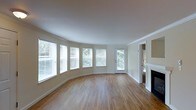 This single-floor space is defined by its graceful architectural details and warm, inviting atmosphere. The living area immediately draws attention with its striking curved wall of windows, fitted with white horizontal blinds, which floods the room with abundant natural light and frames views of surrounding greenery. The smooth hardwood floors throughout lend a clean, classic appeal and complement the neutral, cream-toned walls. Crown molding adds a refined architectural touch, framing the ceiling and reinforcing the space's polished character. A traditional fireplace anchors one end of the room, serving as a natural focal point and adding warmth and charm to the overall aesthetic. A glass-paneled door provides direct access to the outdoors, enhancing the connection to natural surroundings and further contributing to the bright, open feel. The curved layout of the main living area creates a sense of unique spatial flow, distinguishing this space from more conventional floor plans. The combination of generous natural light, warm wood flooring, elegant molding, and the curved windowed wall results in a cohesive, well-appointed space with both character and functionality.