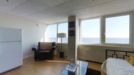 This single-floor space offers an inviting and well-lit atmosphere, defined by its expansive windows that frame breathtaking panoramic views of the open water. The living area is a standout feature, showcasing large horizontal windows along multiple walls that flood the room with natural light and provide unobstructed sightlines to the horizon, creating a serene, coastal ambiance throughout the day.

The space is finished with warm hardwood-style flooring that complements the neutral wall tones and white drop-panel ceiling, lending a clean and cohesive aesthetic. The ceiling height is modest and consistent, adding a sense of coziness while the wide windows counterbalance with an airy openness. A structural column is integrated into the layout, subtly dividing the window arrangements without interrupting the overall flow of the room.

The living area is thoughtfully arranged, with comfortable seating including a classic leather armchair accented with a patterned throw pillow, a floor lamp, and a glass-top coffee table — all contributing to a relaxed yet functional setting. A baseboard heating unit is visible beneath the windows, ensuring comfort year-round. Decorative artwork displayed on the column adds a personal touch to the space, making it feel both livable and characterful.