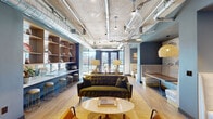 Located at 539 Foster St in the heart of Durham, NC, this single-floor amenity space blends industrial and contemporary design into a cohesive, inviting environment. Exposed concrete ceilings with visible ductwork and pendant lighting establish an urban loft aesthetic, while light wood flooring and muted blue accent walls add warmth and color throughout. The open-plan layout accommodates a variety of uses, anchored by a central lounge area featuring a tufted olive velvet sofa, mid-century-style armchairs, and a round coffee table arranged atop a textured area rug. Adjacent to the lounge, built-in booth seating with white upholstered benches and dark wood tables offers intimate gathering spots beneath woven pendant lights. A sleek wet bar runs along one wall, outfitted with open wood shelving, teal tile backsplash, stainless steel appliances, and a white countertop — flanked by tan bar chairs with gold-tone legs. Floor-to-ceiling windows flood the space with natural light and frame views of the surrounding Durham streetscape and neighboring buildings. A small café-style seating nook positioned directly at the windows provides a bright, airy spot for casual meetings. The space is accessed via a tiled corridor with elevator access, with a posted maximum occupancy of 49 persons.