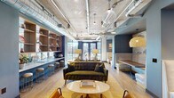 Located at 539 Foster St in the heart of Durham, NC, this single-floor amenity space blends industrial and contemporary design into a cohesive, inviting environment. Exposed concrete ceilings with visible ductwork and pendant lighting establish an urban loft aesthetic, while light wood flooring and muted blue accent walls add warmth and color throughout. The open-plan layout accommodates a variety of uses, anchored by a central lounge area featuring a tufted olive velvet sofa, mid-century-style armchairs, and a round coffee table arranged atop a textured area rug. Adjacent to the lounge, built-in booth seating with white upholstered benches and dark wood tables offers intimate gathering spots beneath woven pendant lights. A sleek wet bar runs along one wall, outfitted with open wood shelving, teal tile backsplash, stainless steel appliances, and a white countertop — flanked by tan bar chairs with gold-tone legs. Floor-to-ceiling windows flood the space with natural light and frame views of the surrounding Durham streetscape and neighboring buildings. A small café-style seating nook positioned directly at the windows provides a bright, airy spot for casual meetings. The space is accessed via a tiled corridor with elevator access, with a posted maximum occupancy of 49 persons.