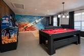 Located at 240 Martin Rd SW in Huntsville, Alabama — home to NASA's Marshall Space Flight Center — this single-floor entertainment space captures the spirit of its surroundings with a bold, space-themed design aesthetic. A striking rocket launch mural dominates one wall of the main game room, complemented by a vivid deep-space painting in the lounge area, setting a cohesive and immersive visual tone throughout.

The game room serves as the centerpiece, featuring a sleek black pool table with a red felt surface, illuminated by an industrial pendant light. Two classic arcade cabinets are displayed alongside a large flat-screen TV, framed by a warm wood-slatted accent wall that adds texture and contrast to the dark color scheme.

Adjacent to the game area is a comfortable lounge zone with a large sectional sofa, tufted cushions, and casual ottomans arranged around a projection screen — ideal for movie screenings or presentations. Multiple windows dressed in blackout curtains allow for flexible lighting control.

A dedicated workstation area offers multiple desks with monitors, making the space functional for both leisure and productivity. The refreshment bar area features industrial pipe shelving, a coffee station, and a butcher-block-topped bar with metal stools — finished with recessed lighting and a jute rug for a modern yet inviting atmosphere.