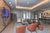 Located at 105 Oak Hill Dr in Athens, Georgia, this single-floor clubhouse space combines contemporary design with warm, inviting finishes to create a refined communal environment. The main lounge serves as the centerpiece, anchored by a striking reclaimed wood accent wall arranged in a geometric chevron pattern and fitted with a wall-mounted flat-screen display. A tray ceiling with integrated ambient uplighting adds architectural depth and a warm glow to the space, complementing the wide-plank hardwood flooring throughout. The seating arrangement is thoughtfully curated, featuring a channeled leather sofa, plush cream armchairs, and vibrant burnt-orange accent chairs gathered around a rustic wood coffee table — creating an atmosphere that is both comfortable and sophisticated. A bar and counter seating area at the rear is illuminated by a row of matte black pendant lights, set against a textured stone tile backsplash, offering an ideal space for casual gatherings. Large black-framed glass doors and windows allow natural light to flow freely through the space while providing views to adjacent areas. A corridor connects to additional rooms, including a carpeted private room with ample natural light, accessible through bold black-framed glass partitions that maintain visual openness while defining each zone. Wall art and decorative accents throughout add character and a locally inspired aesthetic to the overall design.