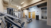 Located at 3031 Norfolk St in Richmond, Virginia, this well-appointed fitness center offers a comprehensive workout environment within a single, open-plan floor. The space features soaring ceilings with exposed structural beams and ductwork, lending an industrial aesthetic that is complemented by bright fluorescent lighting and clean, neutral-toned walls. Durable rubber flooring spans the entire area, providing both safety and functionality for a wide range of exercises. The layout is efficiently organized to accommodate multiple workout zones. A dedicated cardio section houses treadmills, elliptical trainers, and a stationary spin bike, each equipped with individual display screens. Nearby, a strength training area features a multi-station cable machine, a lat pulldown unit, and various resistance machines suited for full-body conditioning. Free weights, including a neatly racked dumbbell set and multiple adjustable benches, occupy the central floor space, allowing for versatile training options. Glass partition walls divide sections of the room, adding a modern architectural detail while maintaining an open, airy feel. At the rear of the space, wood-toned doors lead to restroom facilities, and a water fountain station provides convenient hydration access. A wall-mounted television enhances the cardio area. The overall ambiance is clean, functional, and purpose-built for fitness.