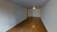 This single-floor space presents a straightforward and functional layout centered around an open main room. The room is finished with warm-toned hardwood flooring that runs continuously throughout, adding a sense of cohesion and warmth to the otherwise neutral interior. The walls are painted in a clean, off-white tone, contributing to a bright and airy atmosphere despite the absence of visible windows in the primary view. Overhead, a flush-mount ceiling light provides direct illumination, keeping the space well-lit. The ceiling is standard height, offering a comfortable and practical sense of enclosure. Toward the far end of the room, two doorways are visible — one appearing to lead to a closet or storage area, and the other serving as an entry or passage to an adjoining room or corridor. White-painted door frames and baseboards provide subtle architectural definition against the neutral walls. The overall layout is open and unobstructed, making the space versatile and adaptable for various uses. The clean, move-in-ready condition of the space highlights its simplicity and potential for personalization.