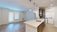 Located at 200 Big Wds Dr in Poquoson, Virginia, this well-appointed one-bedroom, one-bathroom home offers a thoughtfully designed open-concept layout. The combined kitchen and living area serves as the heart of the space, anchored by a peninsula with a crisp white quartz countertop, an undermount sink, and a trio of decorative pendant lights overhead. The kitchen is outfitted with gray shaker-style cabinetry, stainless steel appliances including a refrigerator, range, and microwave, plus open shelving for additional storage. Light wood-look flooring flows throughout the main living areas, lending a clean and contemporary feel. The living room is bright and airy, benefiting from natural light through large windows with blinds and access to an exterior door. The bedroom is a generously sized retreat, carpeted for comfort, with windows that bring in ample natural light. It connects to a spacious walk-in closet equipped with extensive wire shelving and additional carpet underfoot. The full bathroom features a tub-shower combination with brushed nickel fixtures, a vanity, and matching light wood-look flooring. In-unit laundry is conveniently located within the kitchen area, completing this functional and modern home.
