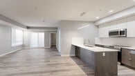 Located in West Garneau, this 2-bedroom, 2-bathroom unit offers a clean, contemporary design across a single floor. Light ash-toned laminate flooring flows seamlessly throughout, complemented by neutral grey walls and crisp white trim that create a bright, cohesive aesthetic.

The kitchen is a standout feature, showcasing a peninsula with a white quartz countertop and an undermount double sink, dark wood-grain lower cabinetry, and a combination of white upper cabinets and dark upper cabinetry. Stainless steel appliances — including a French door refrigerator, slide-in range, and built-in microwave — complete the space, while a sleek linear pendant light adds a modern touch above.

The living area is generously proportioned, with recessed lighting and large windows fitted with roller shades that allow ample natural light to filter in. Both bedrooms are well-sized; one features corner windows and the other includes a double-door closet. Each room maintains the same warm flooring and neutral palette.

The two bathrooms are finished in white marble-look tile with clean, modern chrome fixtures. Both include a tub-shower combination and a vanity with wood-grain cabinetry and a white countertop, with one bathroom featuring a backlit mirror. A hallway connects all rooms, making for an efficient and functional layout throughout.